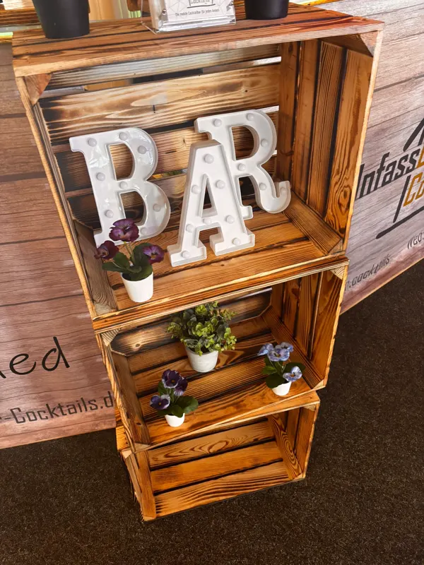 Deko aus Holzkisten mit „BAR“-Schriftzug und Blumen neben der mobilen Bar