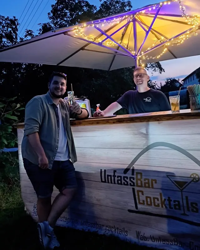 Mobile Bar mit UnfassBar-Cocktails-Banner und beleuchtetem Schirm am Abend