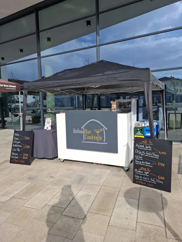 Weiße mobile Bar mit UnfassBar-Logo unter einem Pavillon vor einem Gebäude mit Glasfassade
